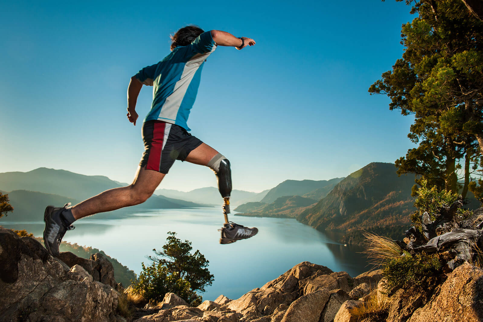 Image of man with prosthetic leg jumping on a mountain with a lake in the background